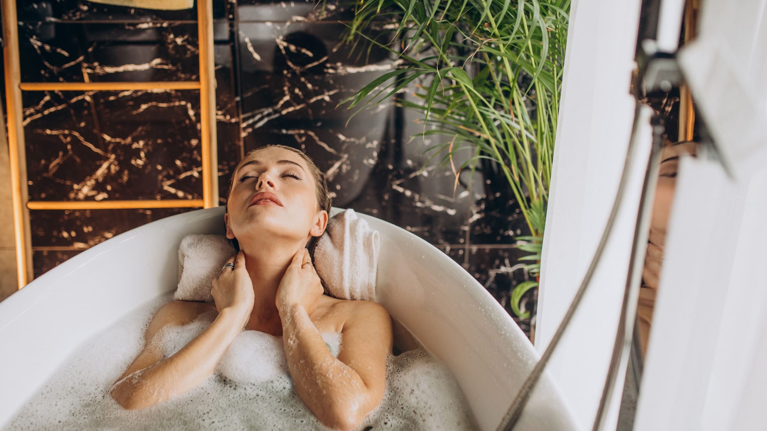 Woman leaning back in a bubble-filled bathtub with towel support and indoor plants creating a calm atmosphere